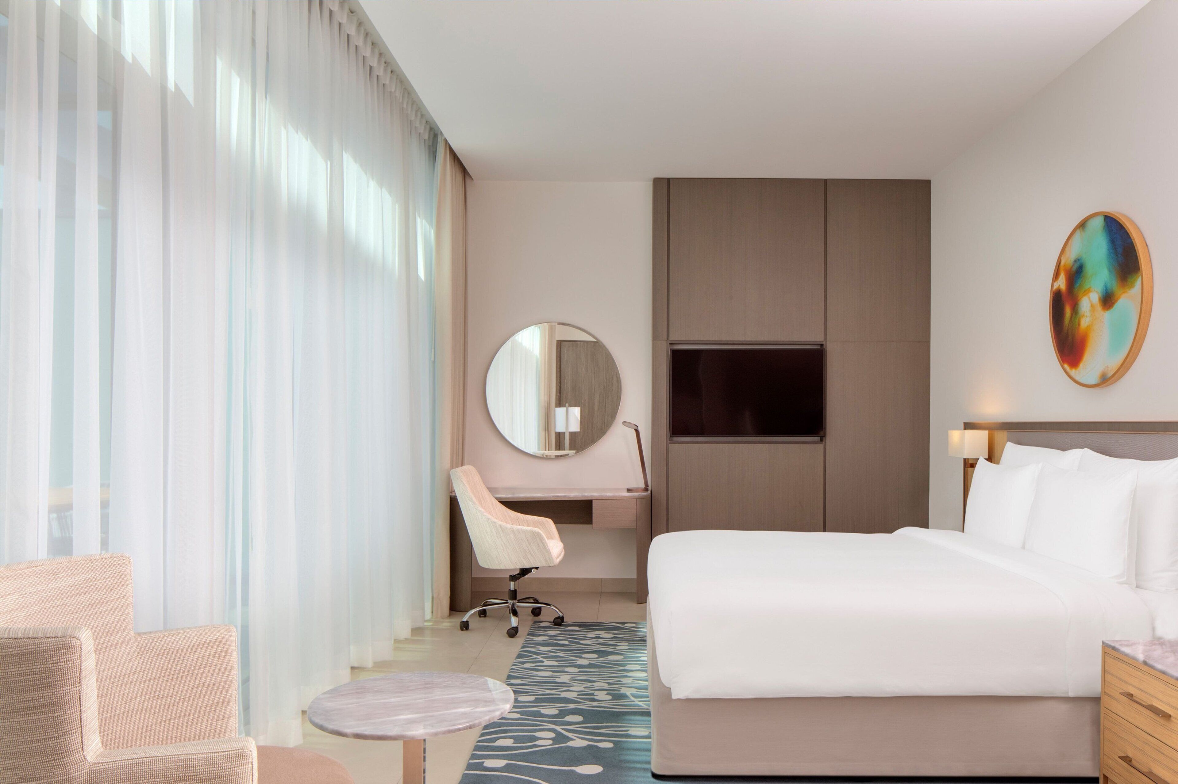 Habitació d'hotel amb un llit, escriptori, cadira, mirall rodó i un quadre a la paret.