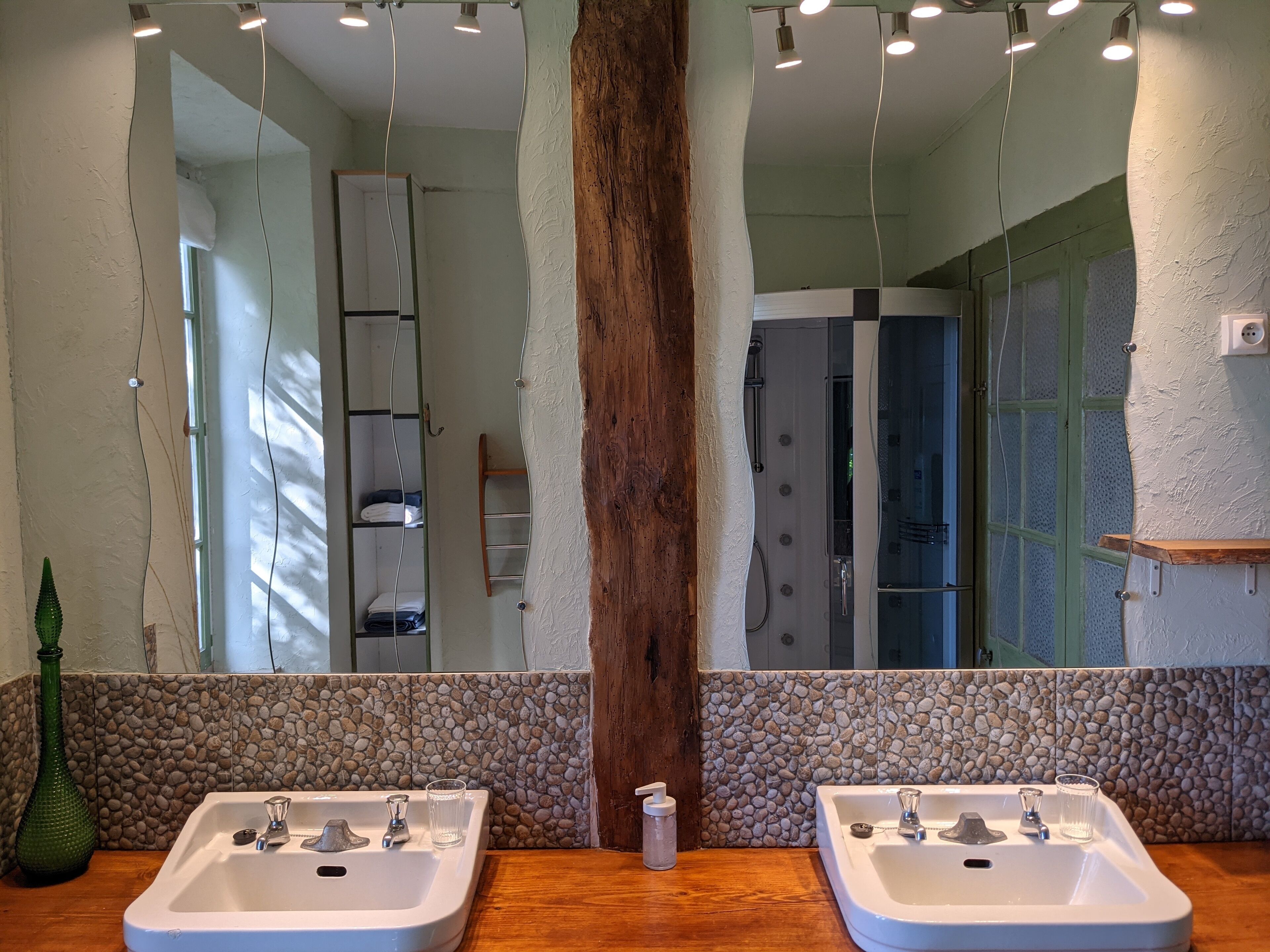 Une salle de bain avec deux lavabos, une colonne en bois et un miroir qui reflète l’espace de la douche.