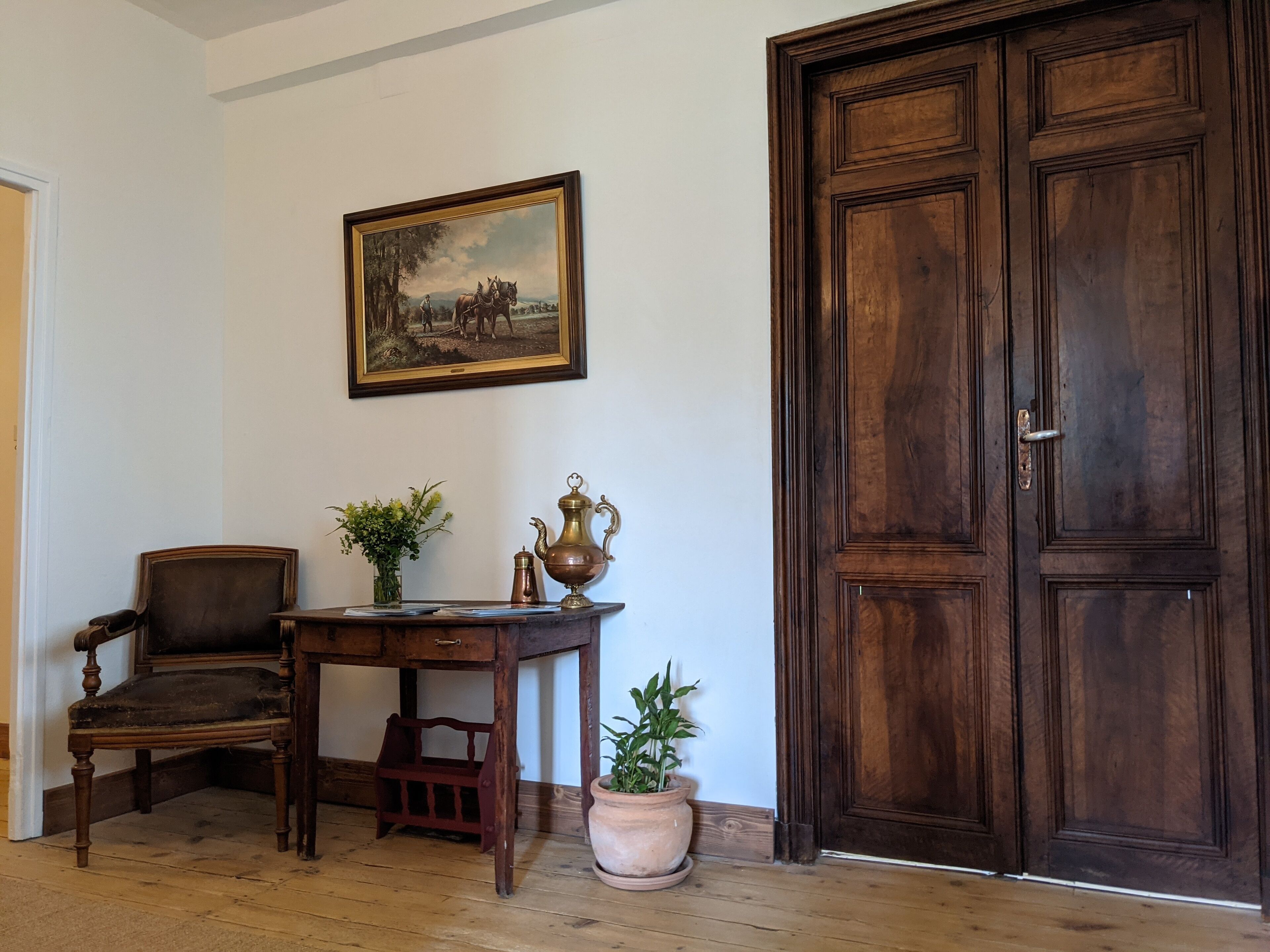 A room with a wooden desk, a chair, a large wooden door, and a framed painting on the wall.