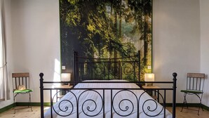 Slaapkamer met een groot wanddecoratie met een bosmotief, een hemelbed, twee nachtkastjes met lampen en een raam met gordijnen.