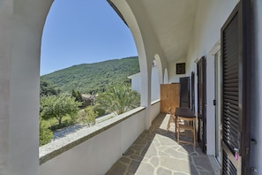 A balcony with a stone floor, wooden chair, and shuttered doors. A view of a green hillside is visible through an archway.