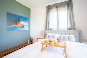 A bedroom with a bed, a bedside table with a tray, a painting on the wall, and a window with curtains.