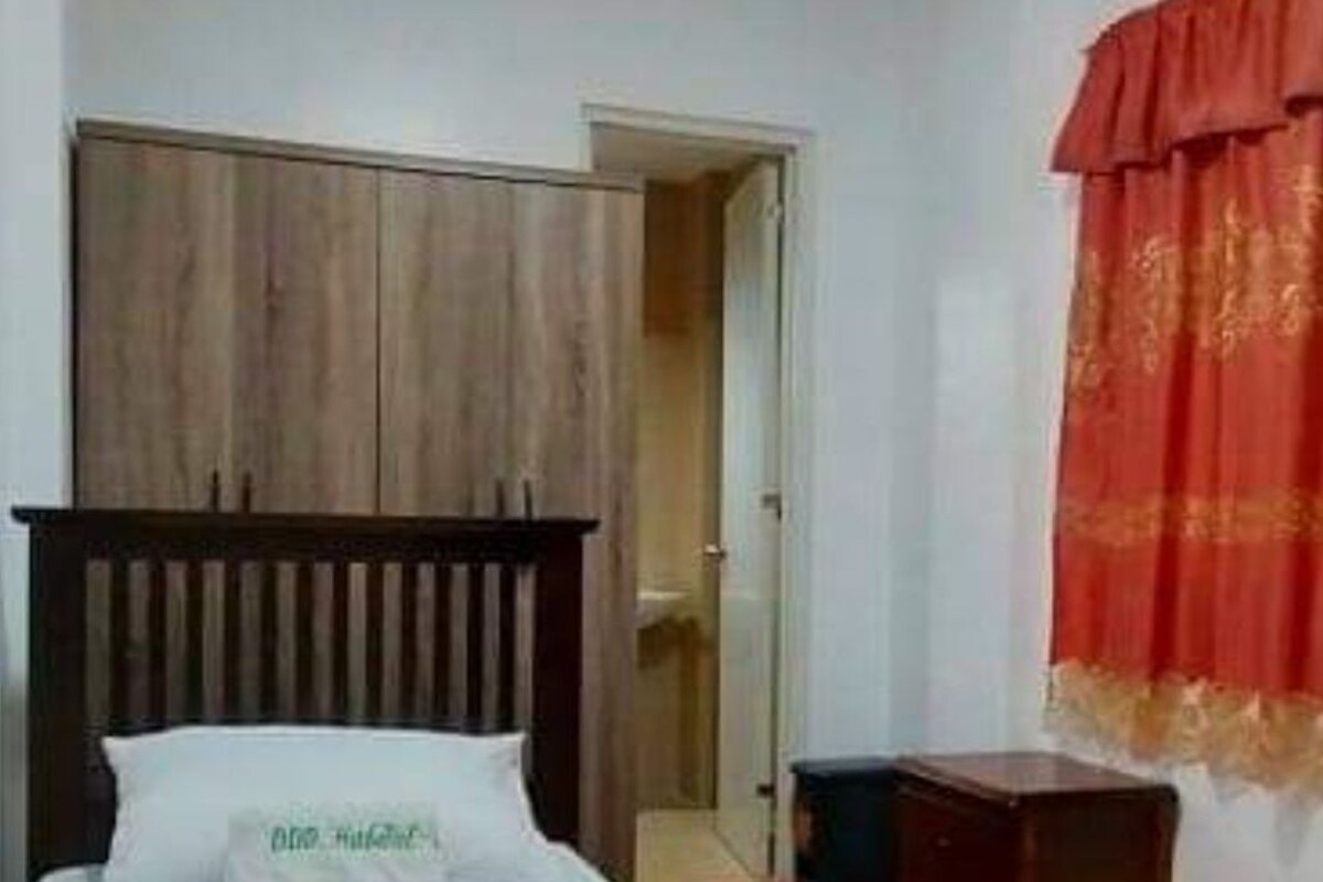 Ein Schlafzimmer mit einem Bett, einem Holzkopfende, einem roten Vorhang und einer kleinen Holzkiste.