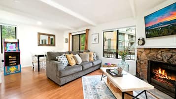 A modern living room with a grey sofa, a fireplace, and a view of the outdoors through large windows.