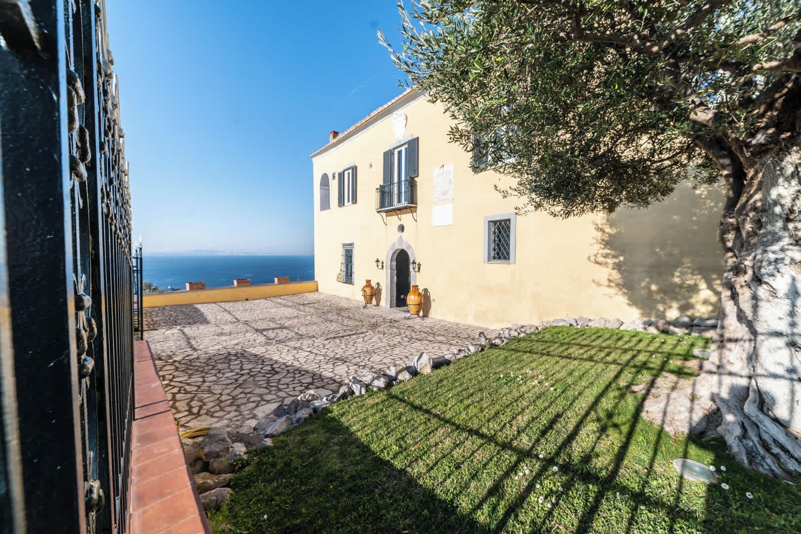 A courtyard with a stone floor, a building with a yellow facade, and a view of the sea.