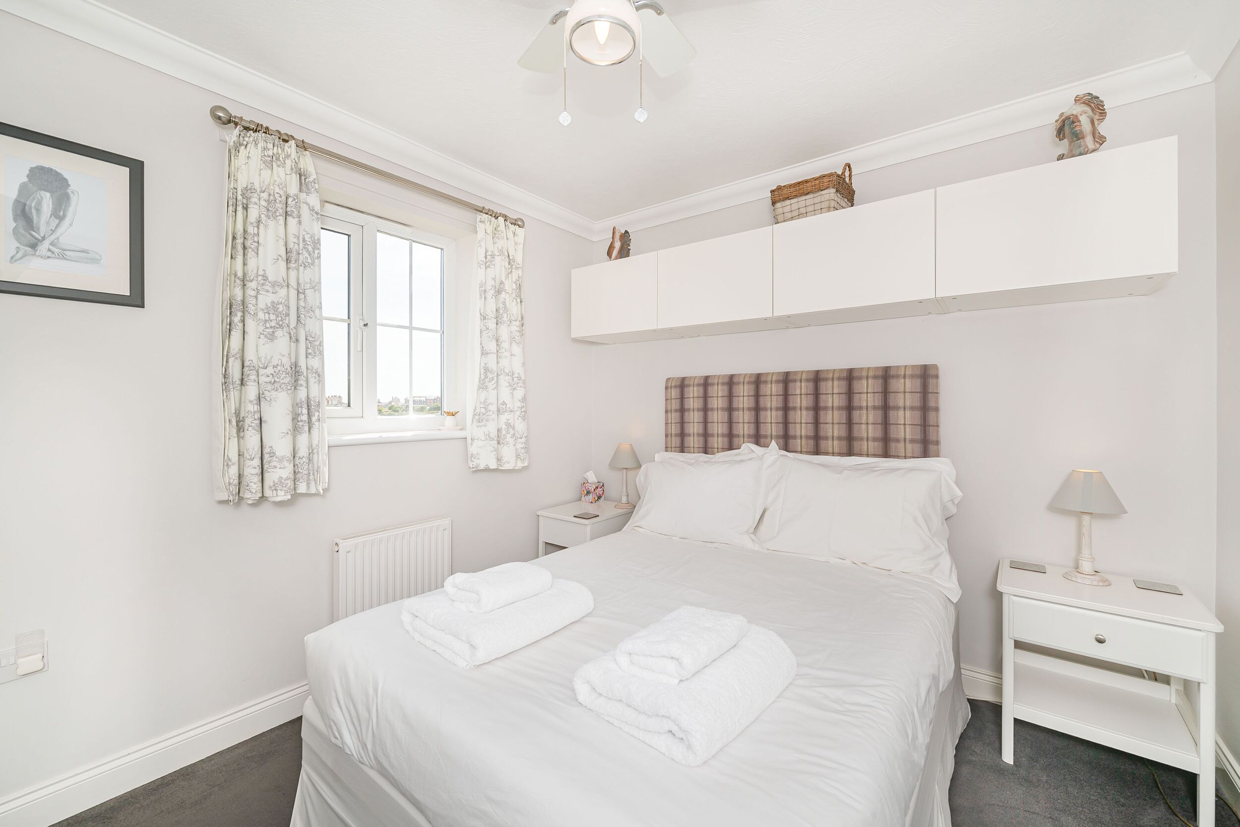 A neatly arranged bedroom with a large bed, a window with curtains, a bedside table with a lamp, and a headboard.