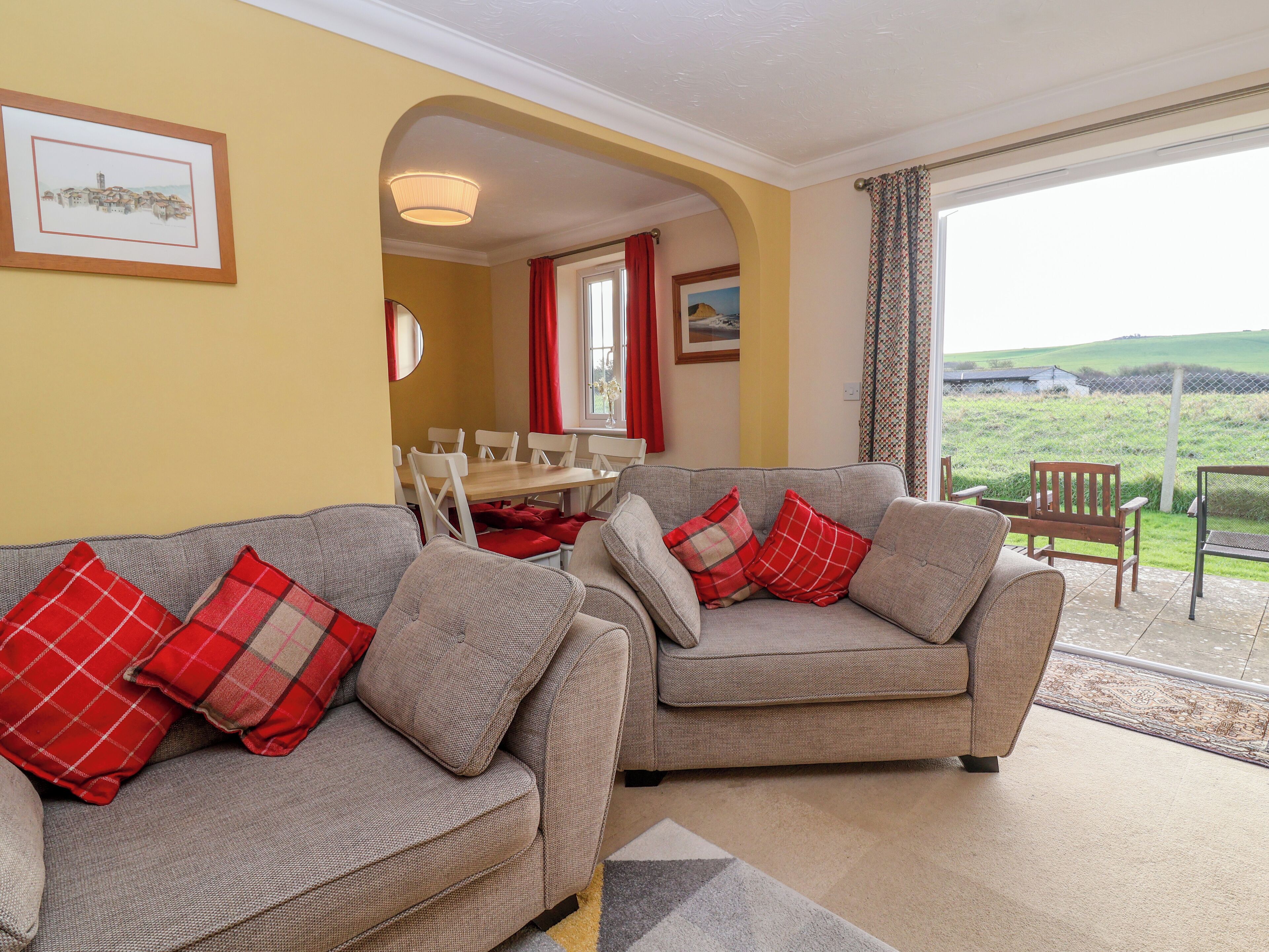 A living room with two grey sofas, red and white checkered cushions, a wooden dining table, and a view of a green landscape through a large window.