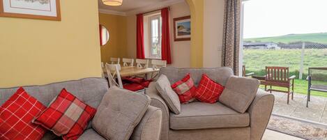 A living room with two grey sofas, red and white checkered cushions, a wooden dining table, and a view of a green landscape through a large window.