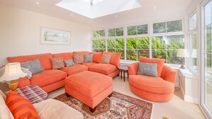 A spacious living room with a large sectional sofa, a round armchair, and a small table. There is a skylight above the sofa and large windows providing a view of greenery outside. - West Bay Holiday Home (Bridport)