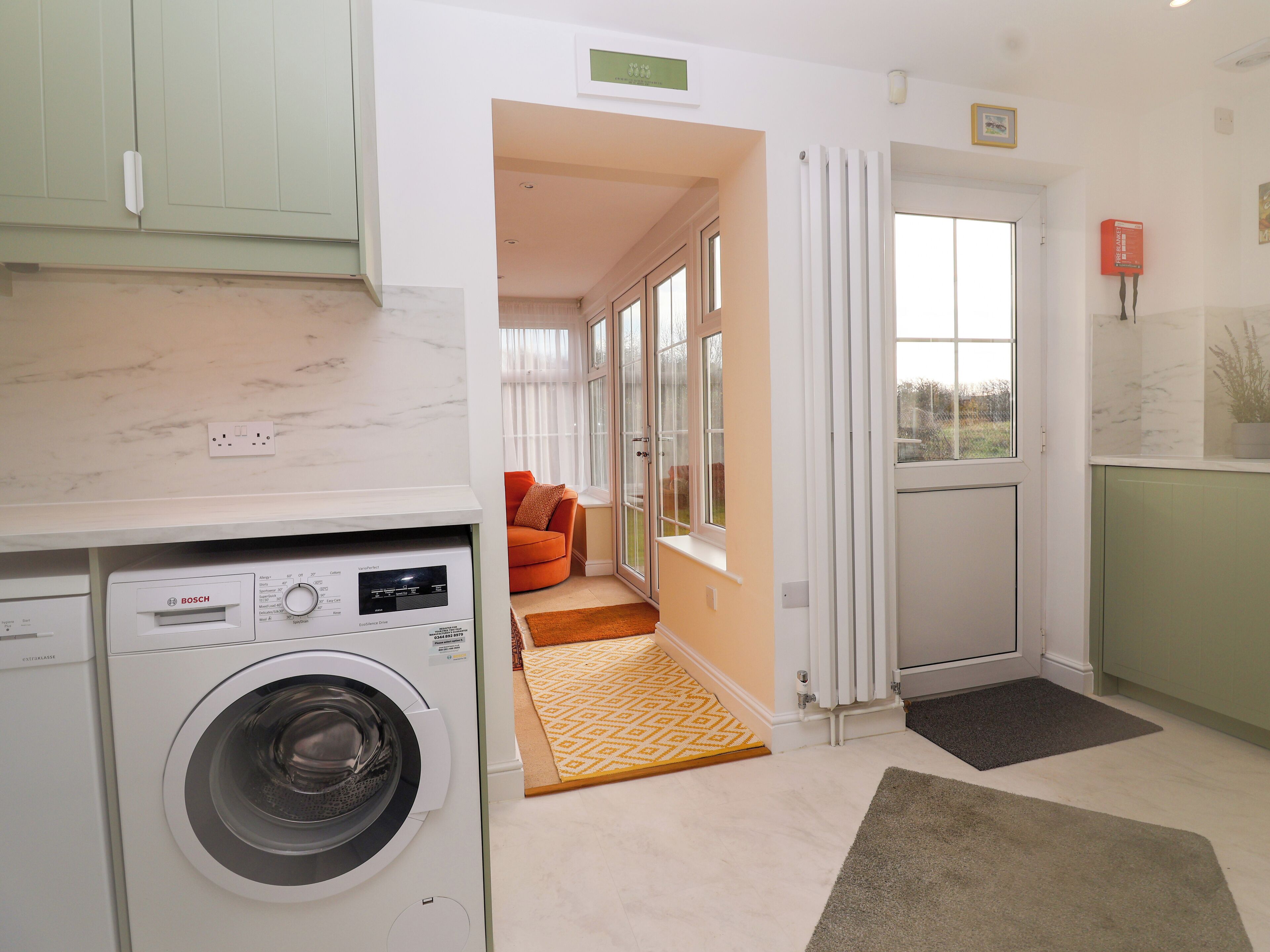 Une petite buanderie équipée d’un lave-linge Bosch, un petit coin salon avec un coussin, et une vue sur l’extérieur à travers une porte vitrée.