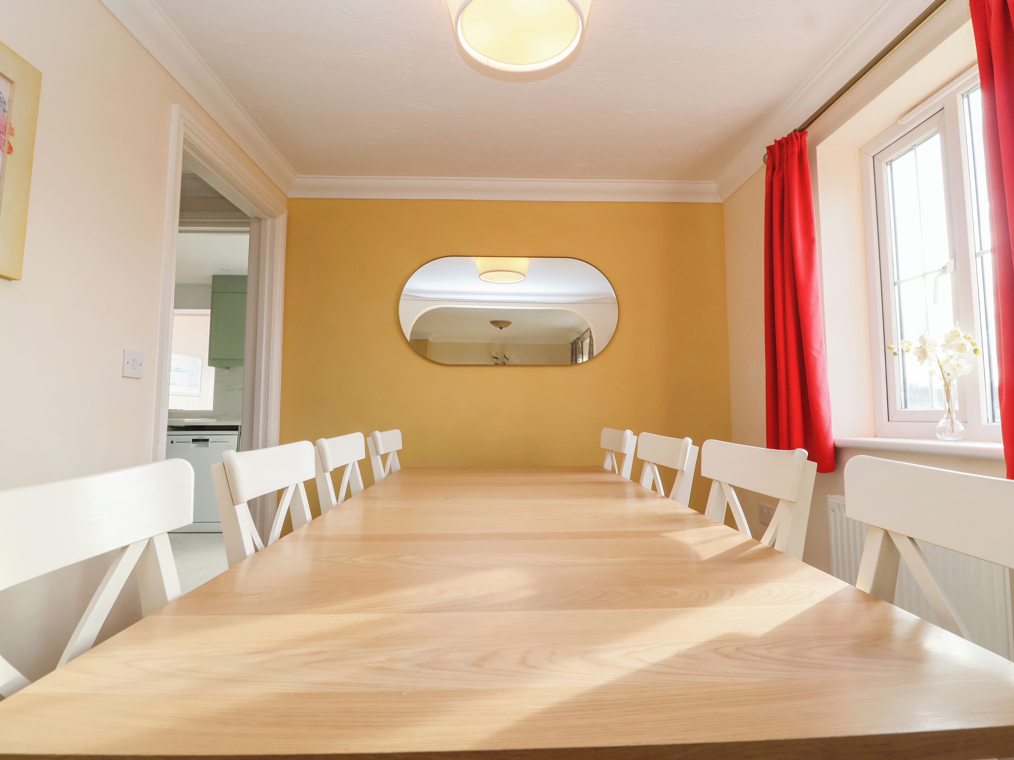 Un comedor con una mesa de madera larga y sillas blancas, una pared de acento amarilla y un espejo grande sobre la mesa.