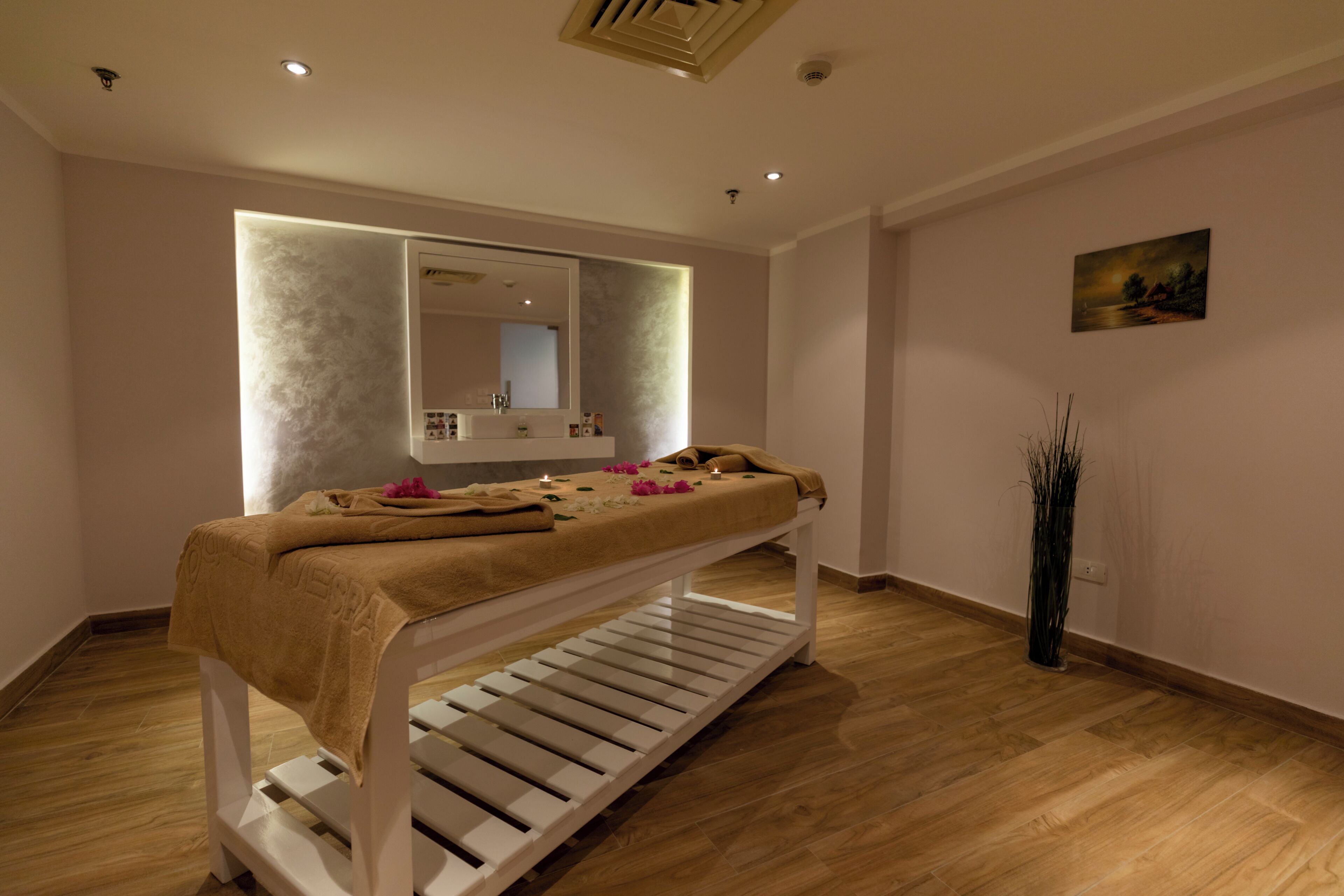 A room with a massage table, a mirror, a painting, and a tall vase with plants.