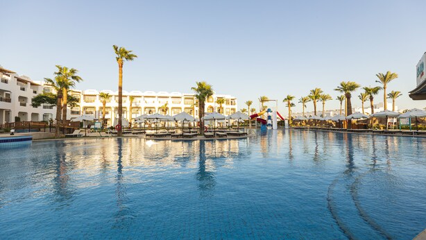 A swimming pool with palm trees and umbrellas, surrounded by hotel buildings.