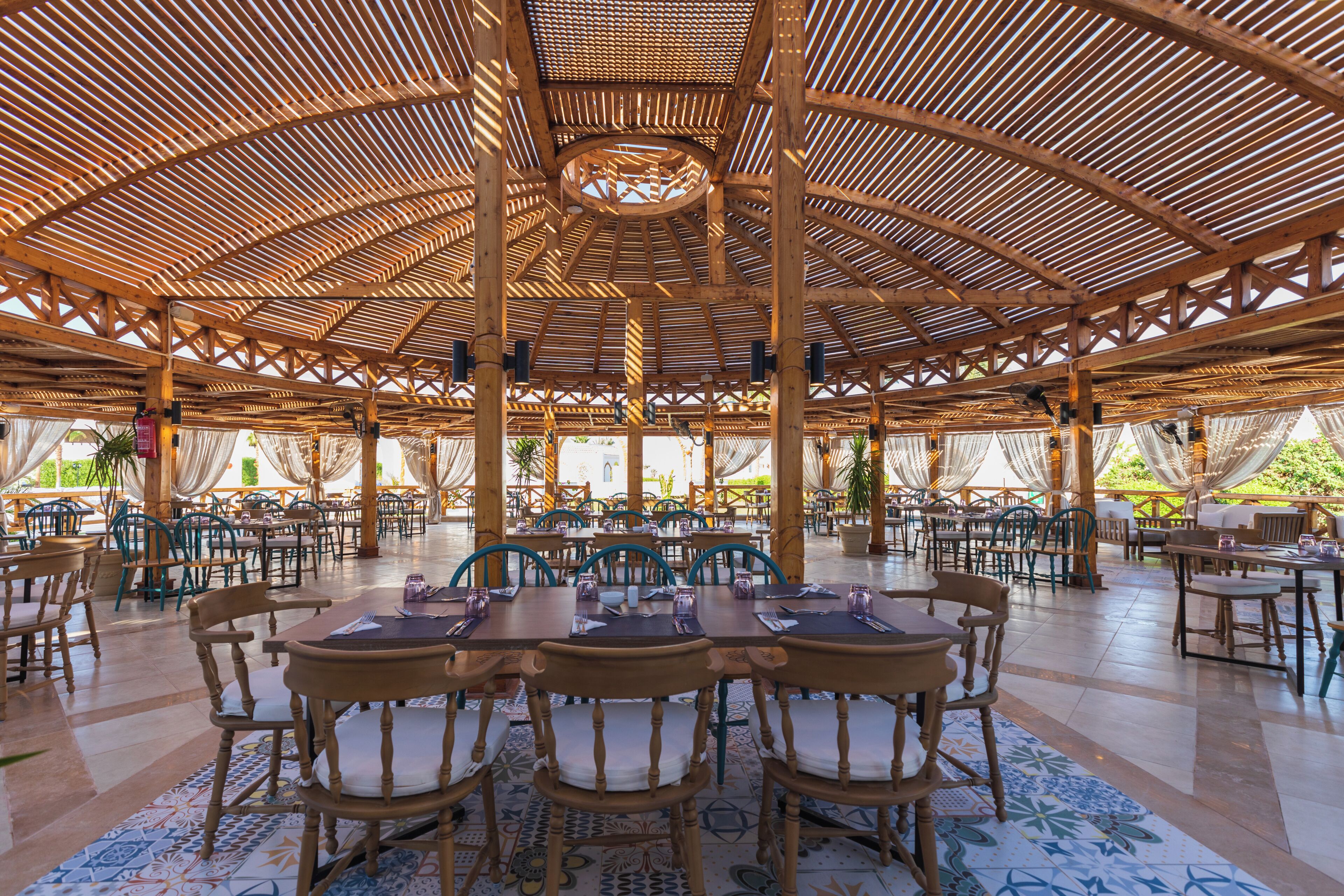 An open-air dining area with wooden tables and chairs, a high ceiling with a wooden lattice design, and a view of the outdoors.