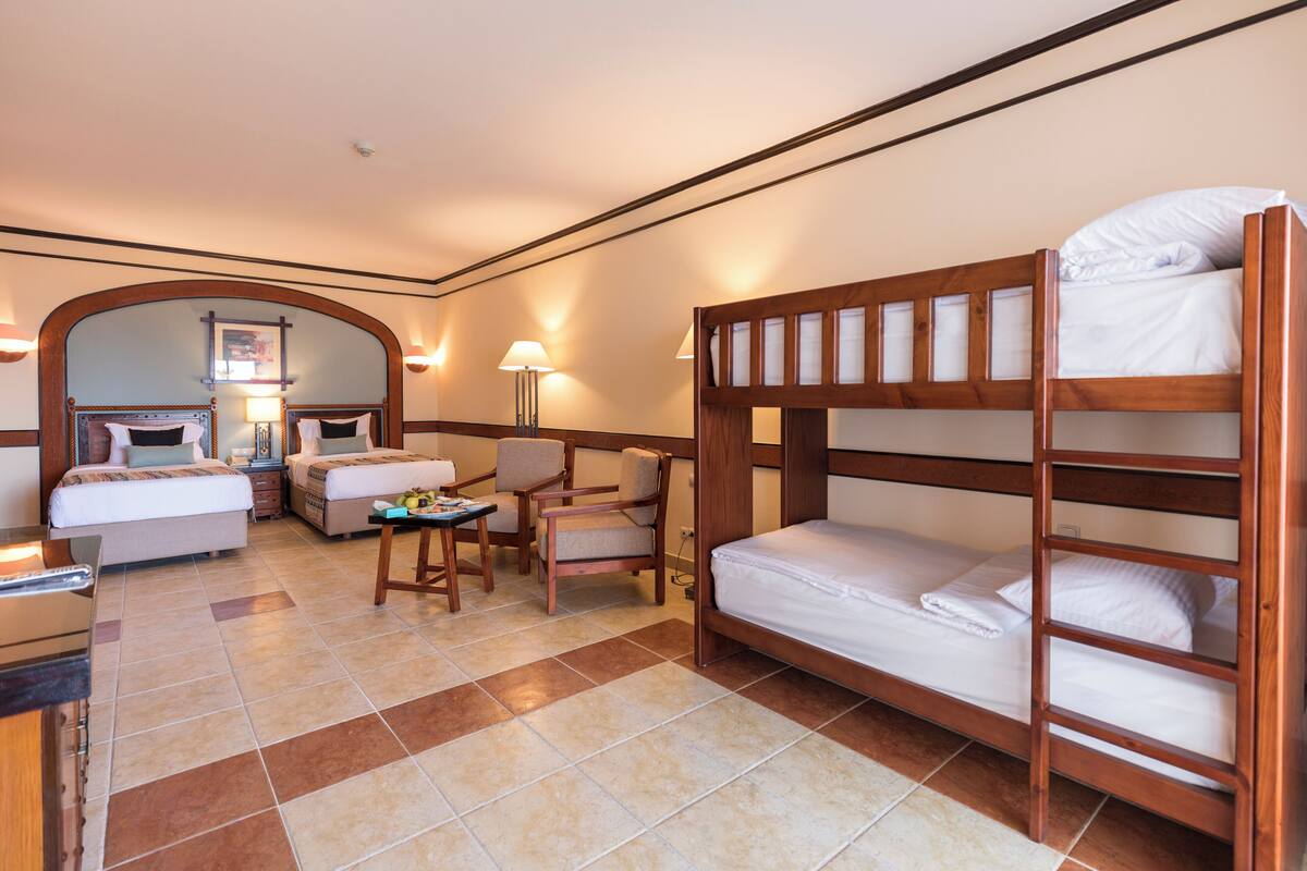 A hotel room with a bunk bed, a desk with a chair, a small table with a lamp, and a bed with a nightstand.