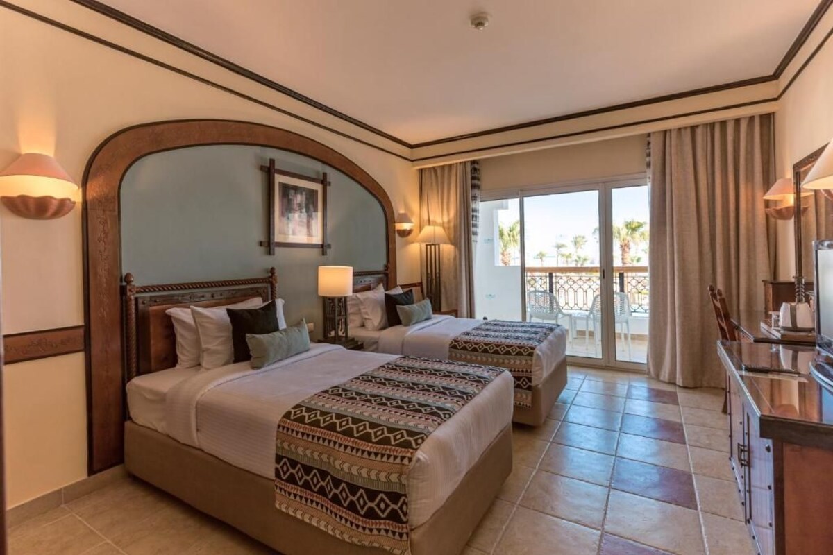 A hotel room with two beds, a balcony, and a view of palm trees.