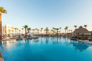 Et resortområde med pool, flere liggestole og parasoller, omgivet af palmer og en klar himmel.