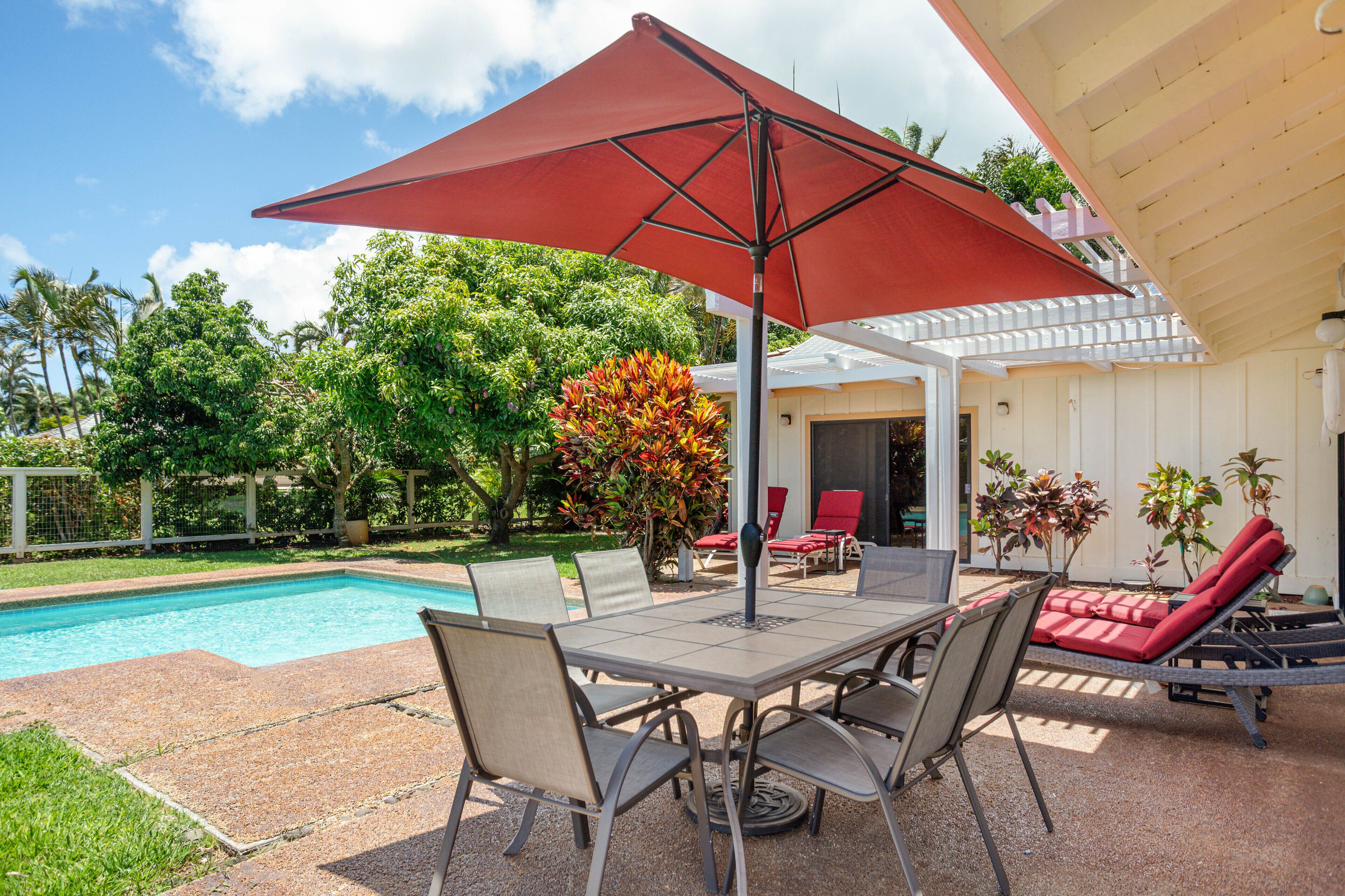 A poolside area with a table and chairs, a red umbrella, and lounge chairs.