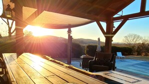 A wooden table with a glass of iced tea under a shaded area, overlooking a pool and outdoor seating. - Cottage on quiet farm close to town and wineries  (Charlottesville)