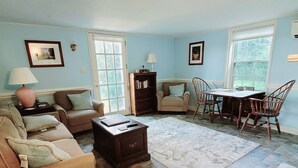 A living room with a sofa, armchair, coffee table, and a dining table with chairs. There is a bookshelf, a lamp, and a trash can. - Cottage on quiet farm close to town and wineries  (Charlottesville)