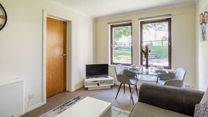 Sala de estar com televisão, duas cadeiras, mesa de centro e sofá. Há duas janelas com vista para um jardim e um edifício.