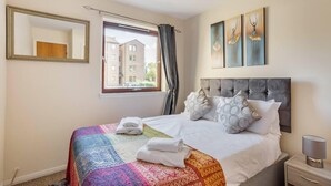 Quarto de hotel com uma cama grande, janela com cortinas, mesa de cabeceira com abajur e obras de arte na parede.