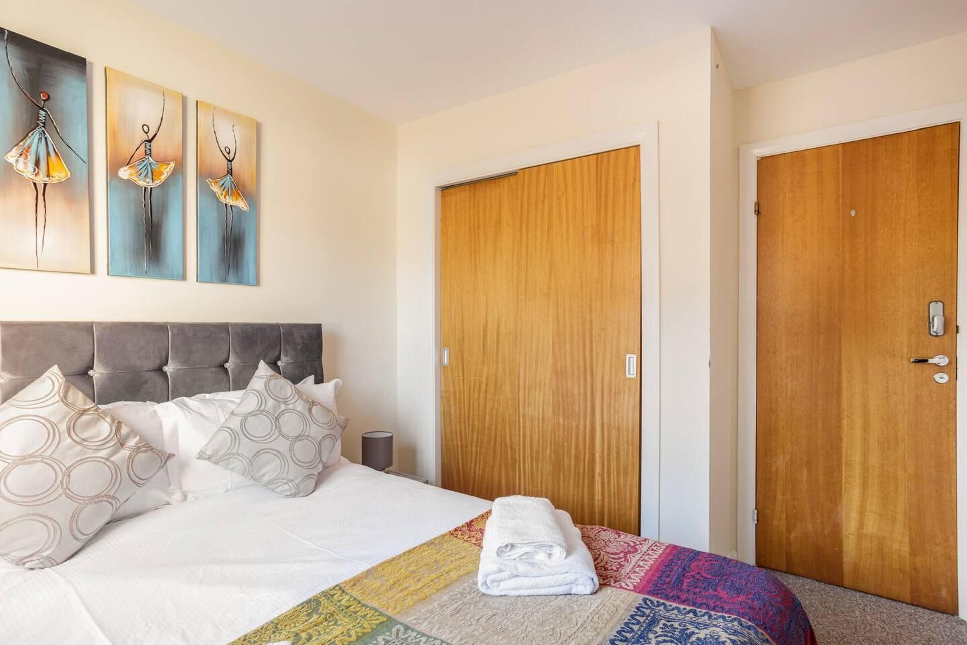 A hotel room with a bed, a set of three paintings, two wooden doors, and a folded towel on the bed.