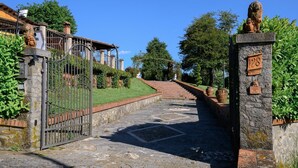 A stone pathway leading to a gated entrance with a lion statue, a stone pillar with a plaque, and a building with a tiled roof in the background. - Colle Santa Cristina, Castelnuovo di Garfagnana, Lucca and Viareggio (Castelnuovo di Garfagnana)
