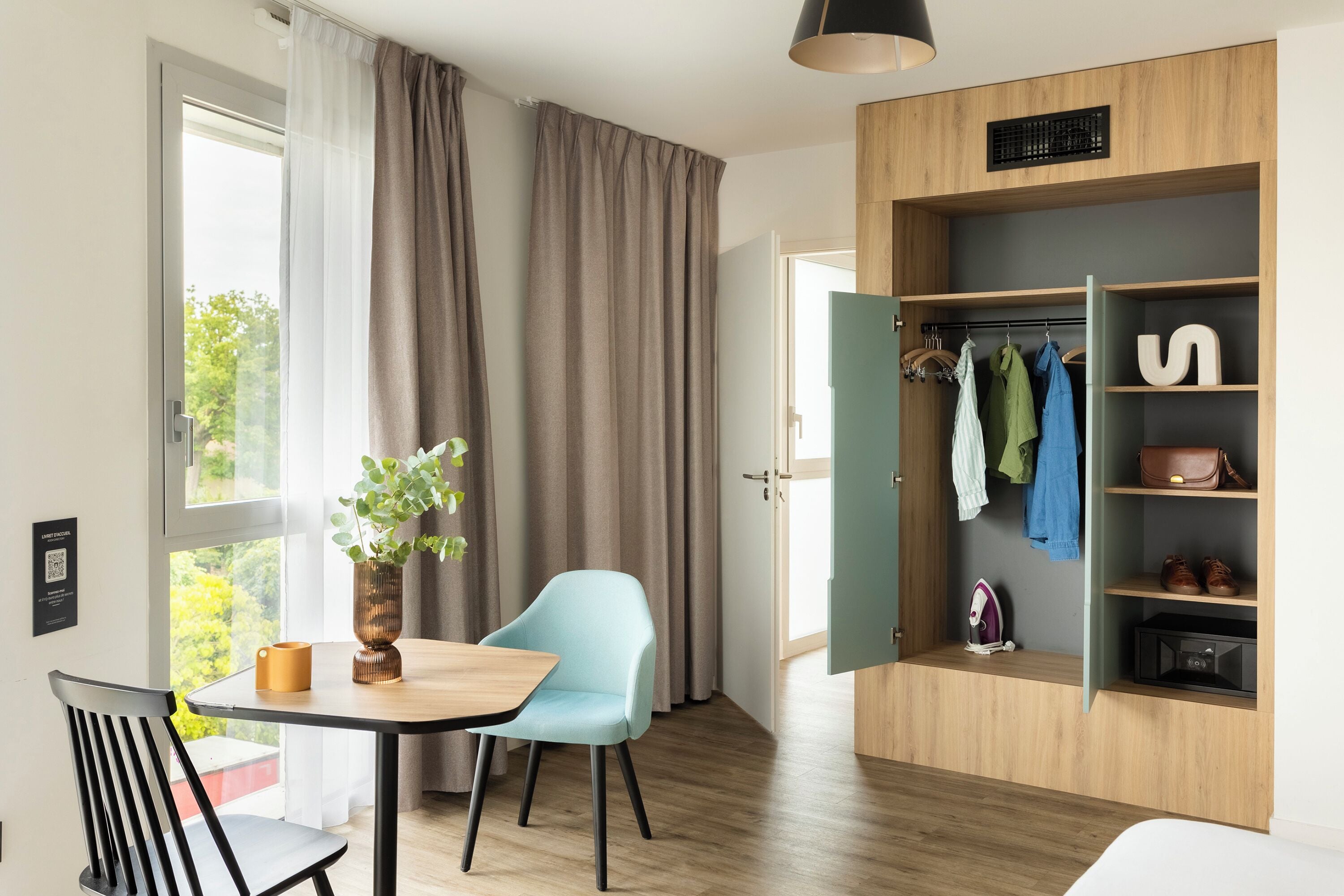 A modern room with a dining table, a chair, a plant, a wardrobe with hanging clothes, and a hanging lamp.