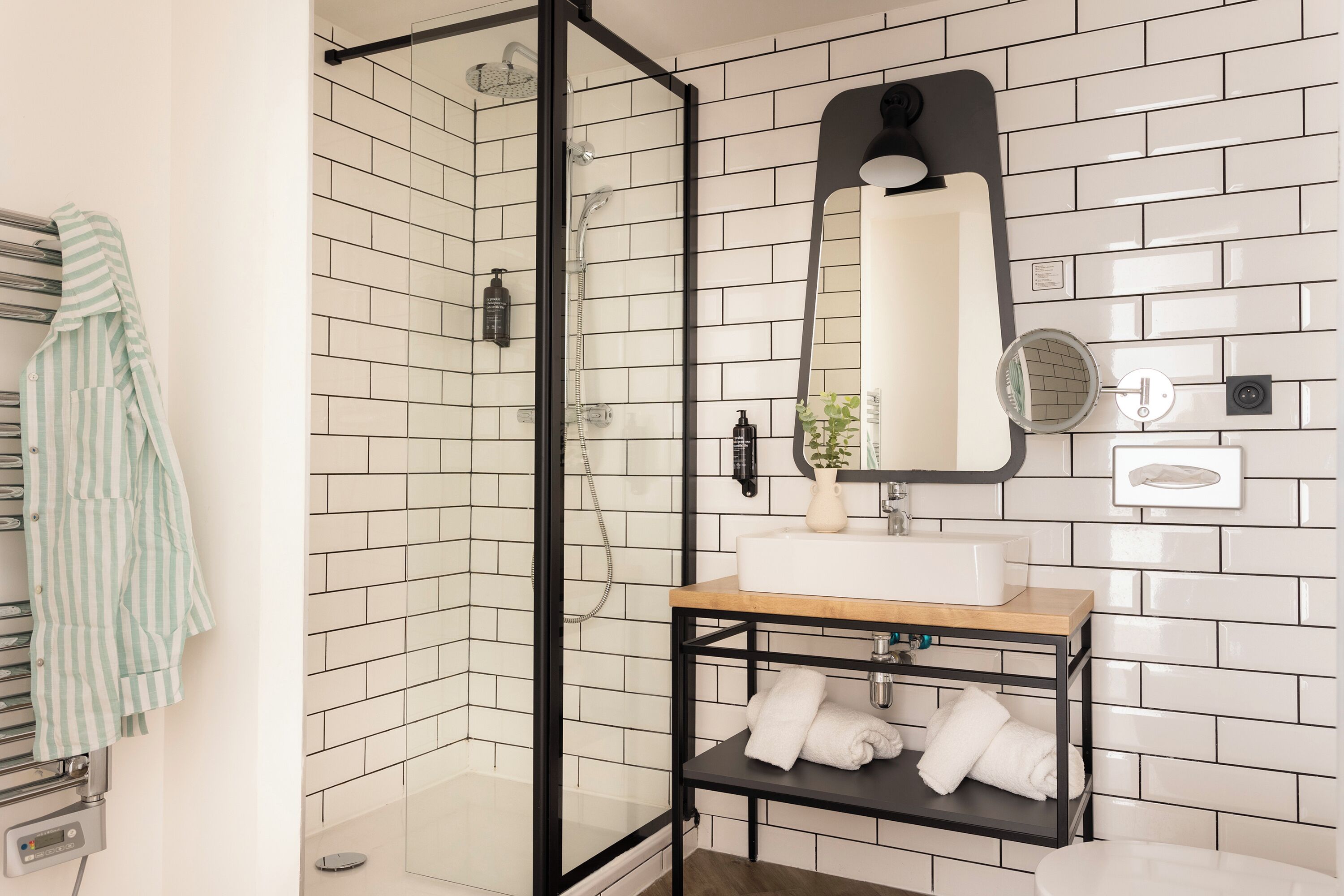 Une salle de bain avec une douche à parois de verre, un lavabo doté d’un plan de travail en bois et une étagère en métal noir où sont rangées les serviettes.