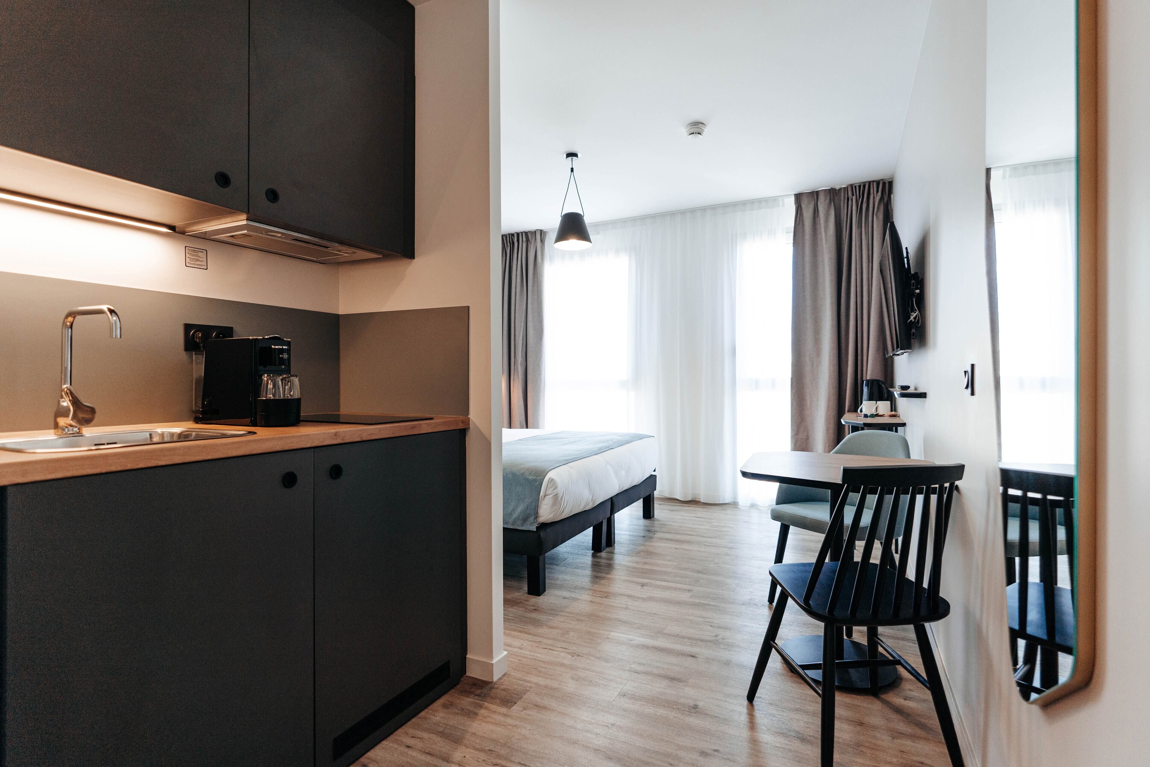 A compact hotel room with a kitchenette, a bed, a dining area, and a large window with curtains.