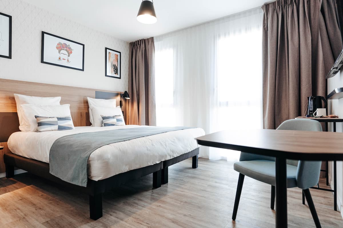 A modern hotel room with a large bed, a bedside table, a desk with a chair, and a window with curtains.