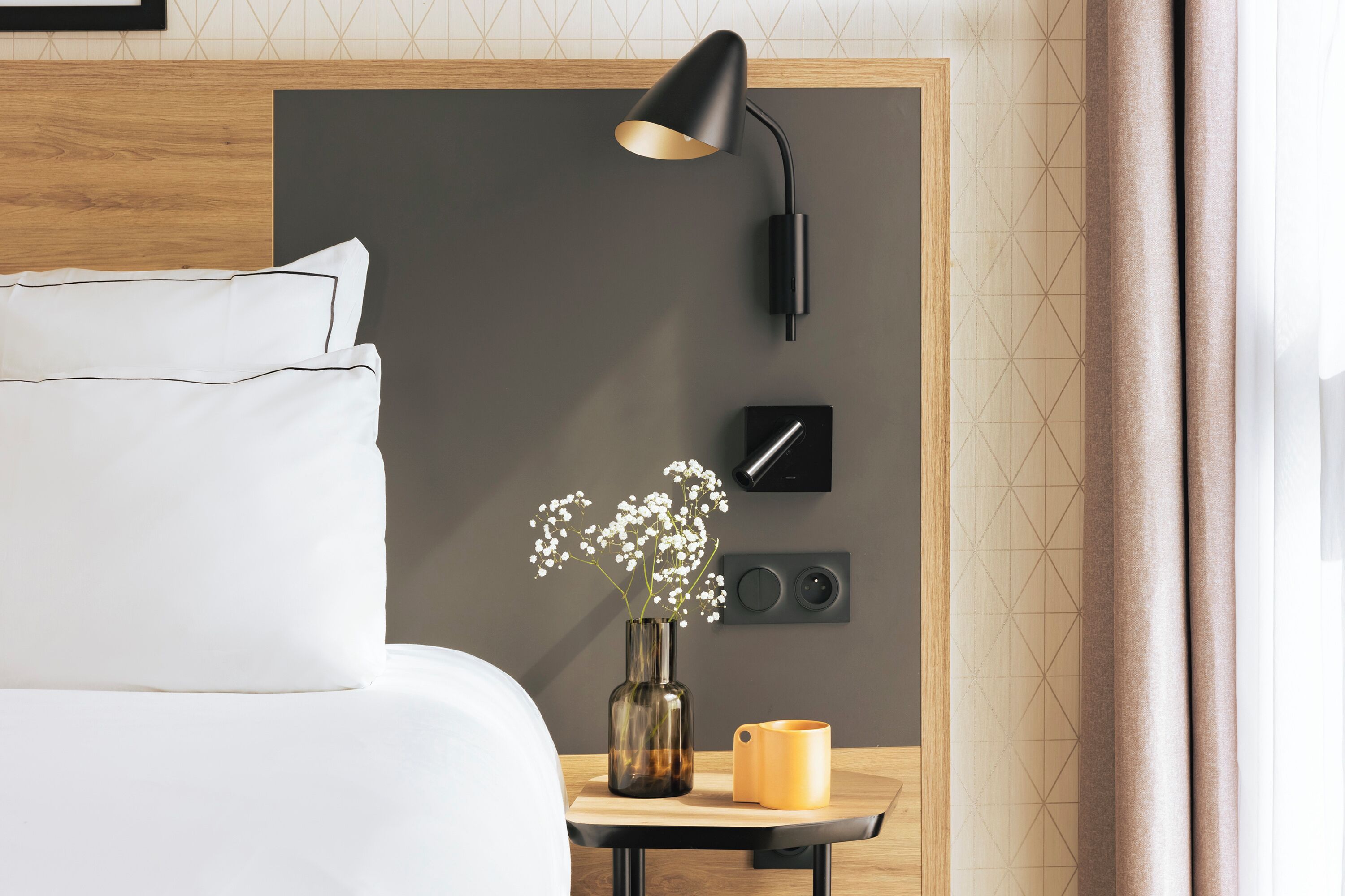 Un lit bien fait, avec du linge de lit blanc, une table de chevet ornée d’un vase de fleurs, une tasse jaune et une lampe fixée au mur.