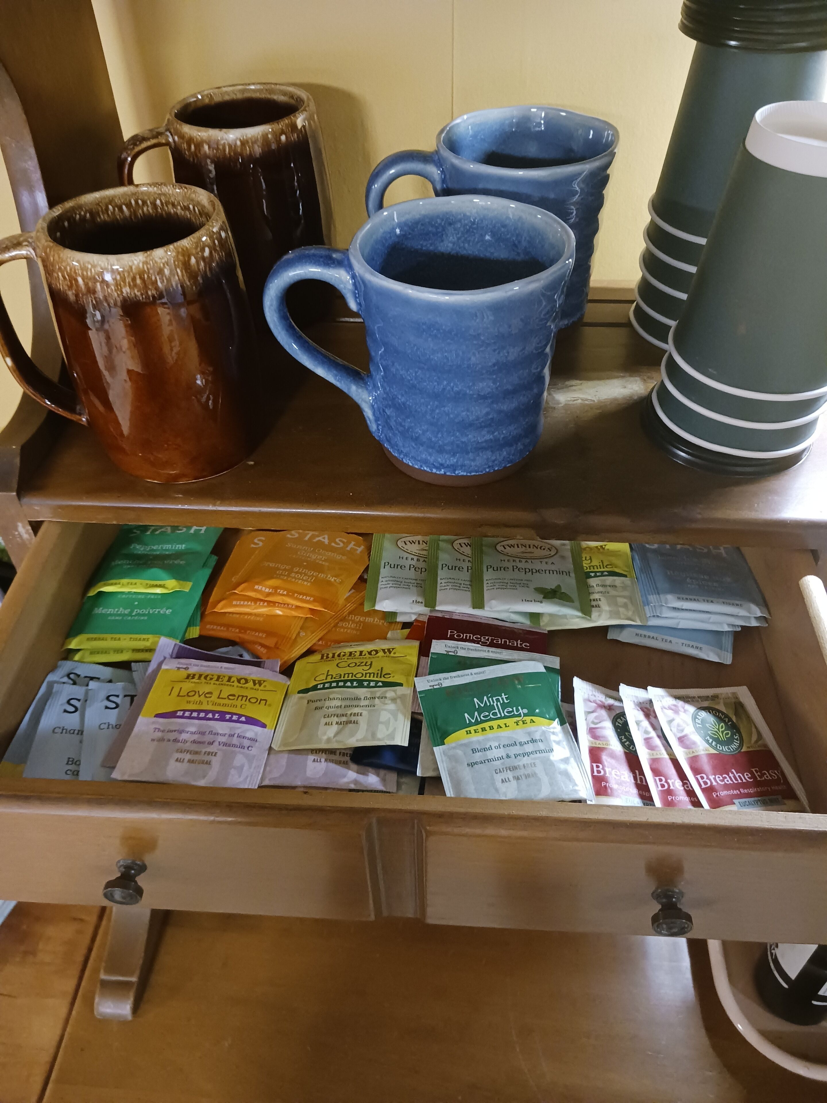 A drawer filled with various herbal tea bags and a collection of mugs on a shelf.