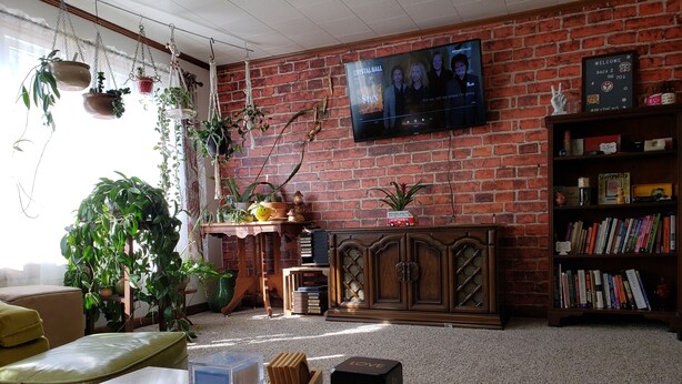 Ein gemütliches Wohnzimmer mit Backsteinwand, einem an einer Holzkommode befestigten Fernseher und einem Bücherregal voller Bücher.