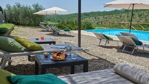 Un espace extérieur avec piscine, des chaises longues et des parasols, entouré d’un sol recouvert de petits galets et de verdure.