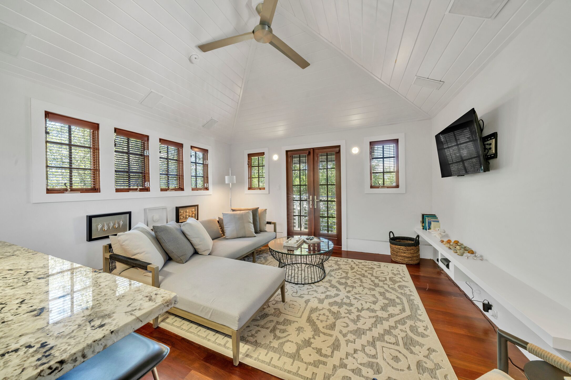 A spacious living area with a sofa, coffee table, and a dining table. There are multiple windows with blinds, a ceiling fan, and a mounted television.