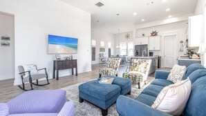 Een ruime woonkamer met een blauwe bank, voetenbankje en schommelstoel, een eethoek met tafel en stoelen, en een keuken met witte kasten en een centraal eiland.