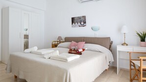 A neatly made bed with a white bedspread and pillows, a bedside table with a lamp, a small table with a plant, and a wall-mounted artwork. - Sunset Home with sea view (Chania)