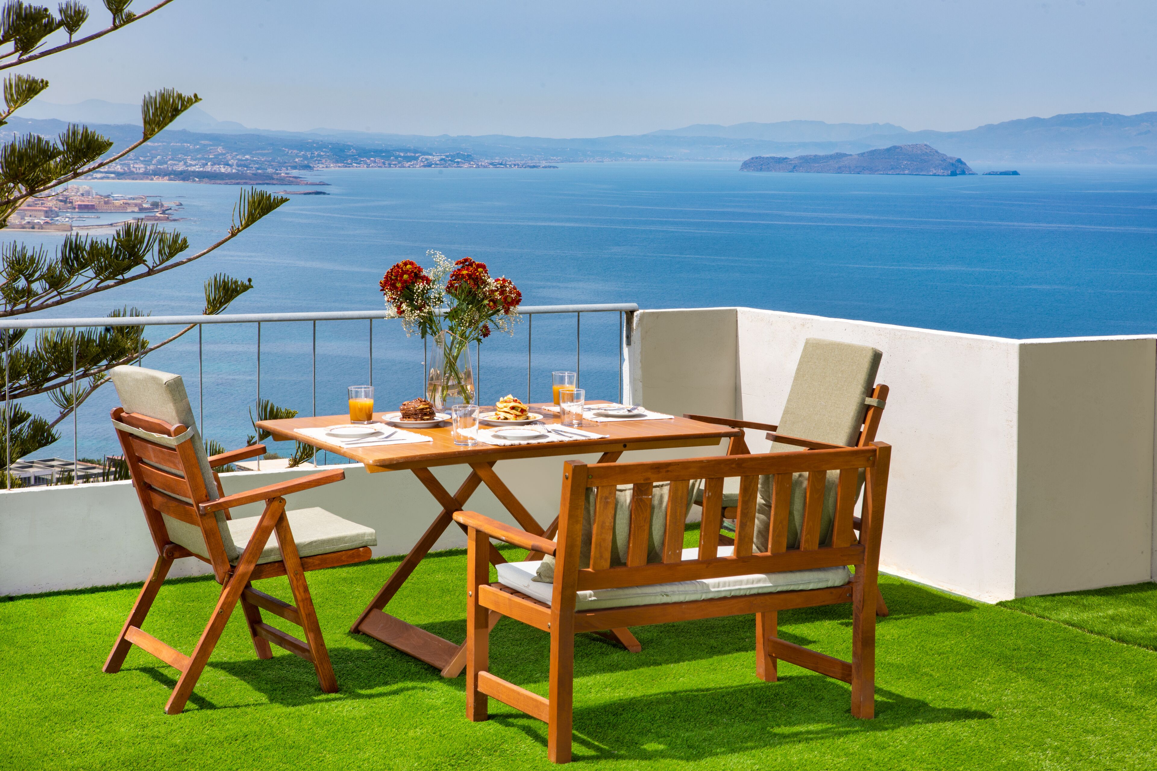 A wooden table and chairs set on a balcony with a view of the sea and distant land.