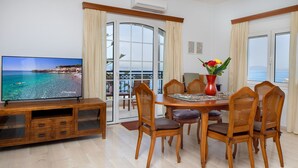A dining area with a wooden table, wicker chairs, a red vase with flowers, and a television displaying a coastal scene. - Sunset Home with sea view (Chania)