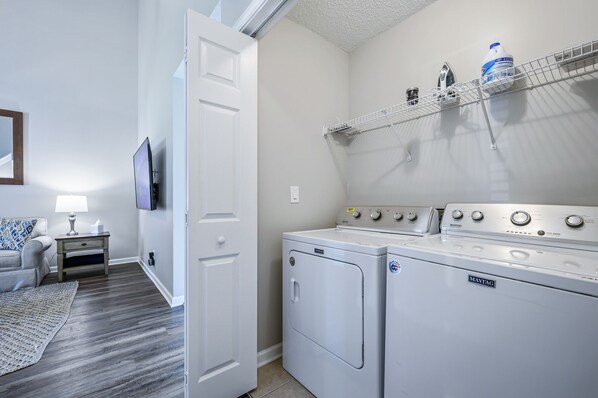Uma lavanderia com duas máquinas de lavar e secar Maytag, uma prateleira com produtos de lavagem, uma televisão e uma pequena mesa com abajur.