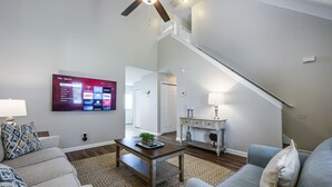 Una sala de estar con un televisor de pantalla plana montado en la pared, una mesa de centro de madera, un sofá y una escalera que conduce a otro nivel.