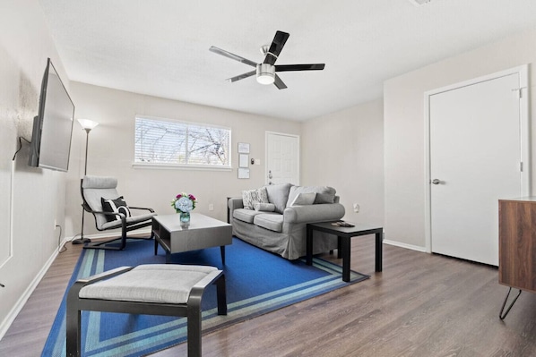 A living room with a sofa, a chair, a coffee table, a TV, a ceiling fan, and a door.