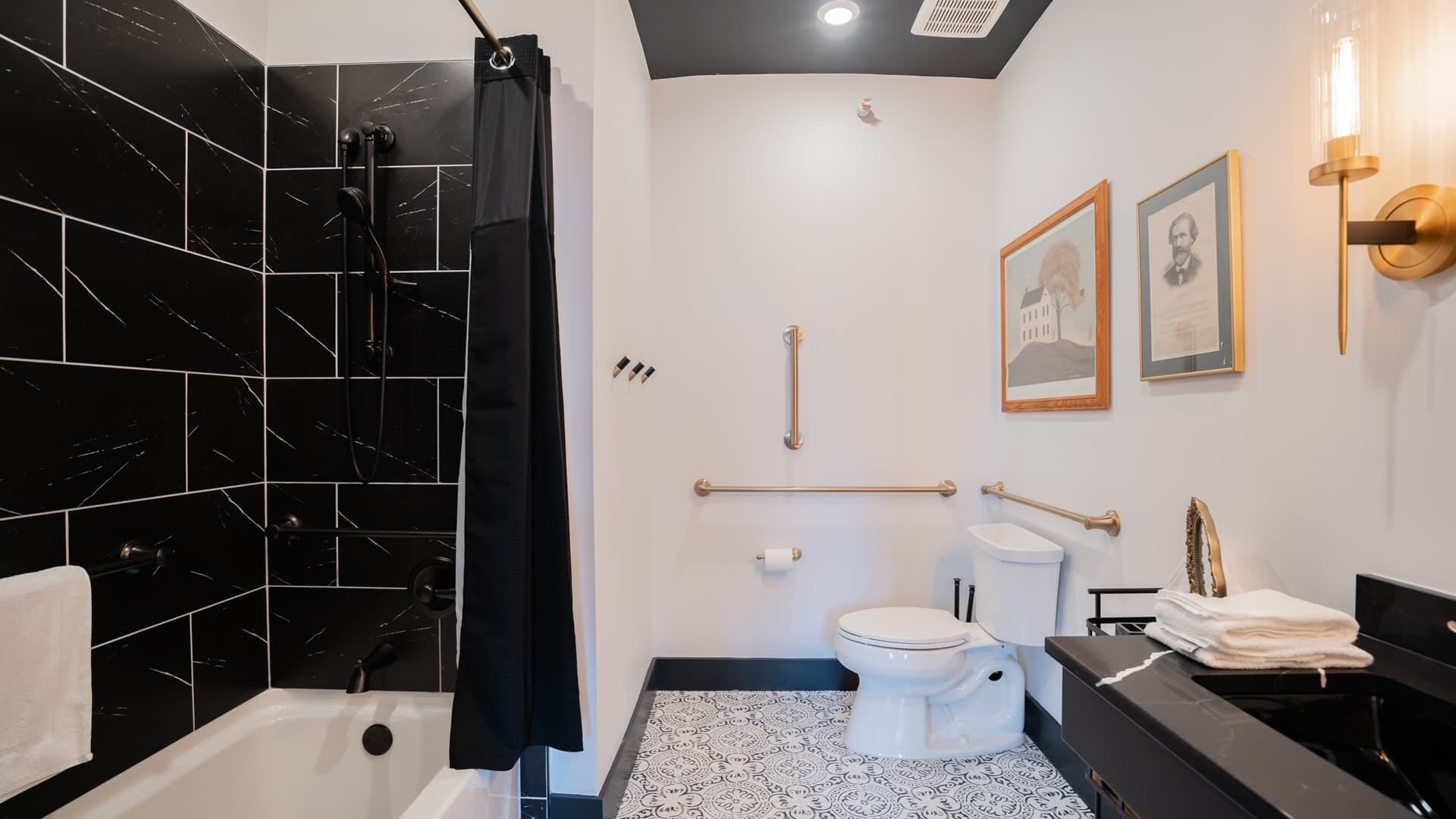 Un baño moderno con zona de ducha revestida de azulejos negros, inodoro blanco y lavabo negro con detalles dorados.
