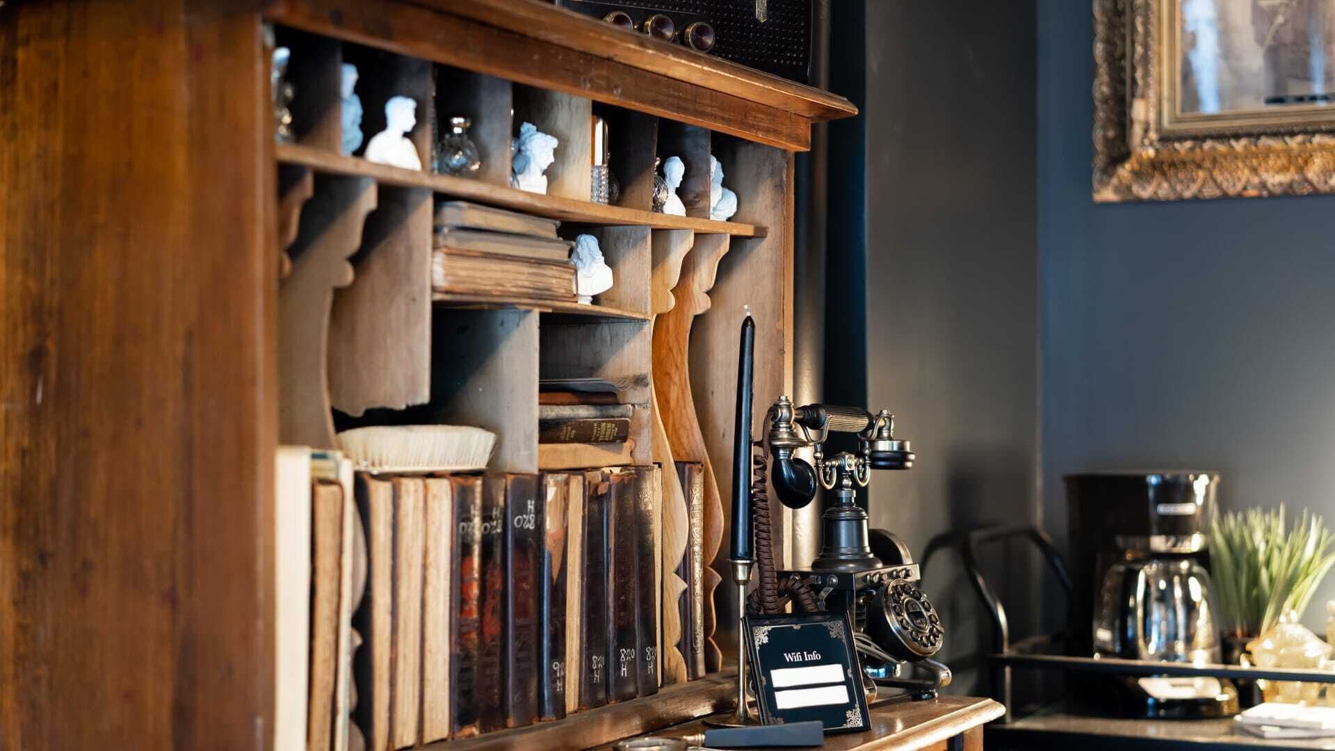 Una estantería de madera llena de libros y bustos decorativos, un teléfono rotatorio antiguo y una pequeña señalización con la palabra “WiFi” sobre una encimera.