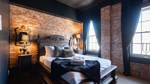 Ein Schlafzimmer mit Backsteinwand, einem großen Bett mit vielen Kissen, einem Nachttisch mit Lampe und einem Fenster mit Vorhängen.