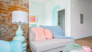 Un salon avec un mur en briques, un canapé de couleur claire, un coussin rose moelleux, une couverture tricotée et une lampe de couleur turquoise.
