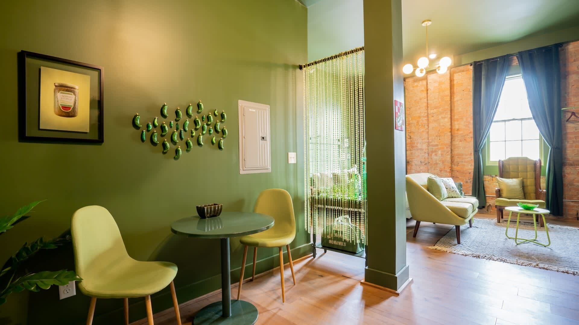 Uma sala de estar aconchegante com uma parede verde, uma pequena mesa redonda, duas cadeiras amarelas e um objeto decorativo na parede.