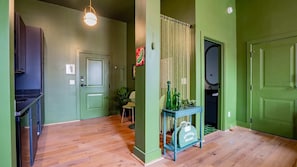 A hallway with green walls, wooden flooring, a table with bottles and glasses, and a door with a mirror. - Pickle Factory Hotel—2BR Near Stadiums & Downtown (Covington)